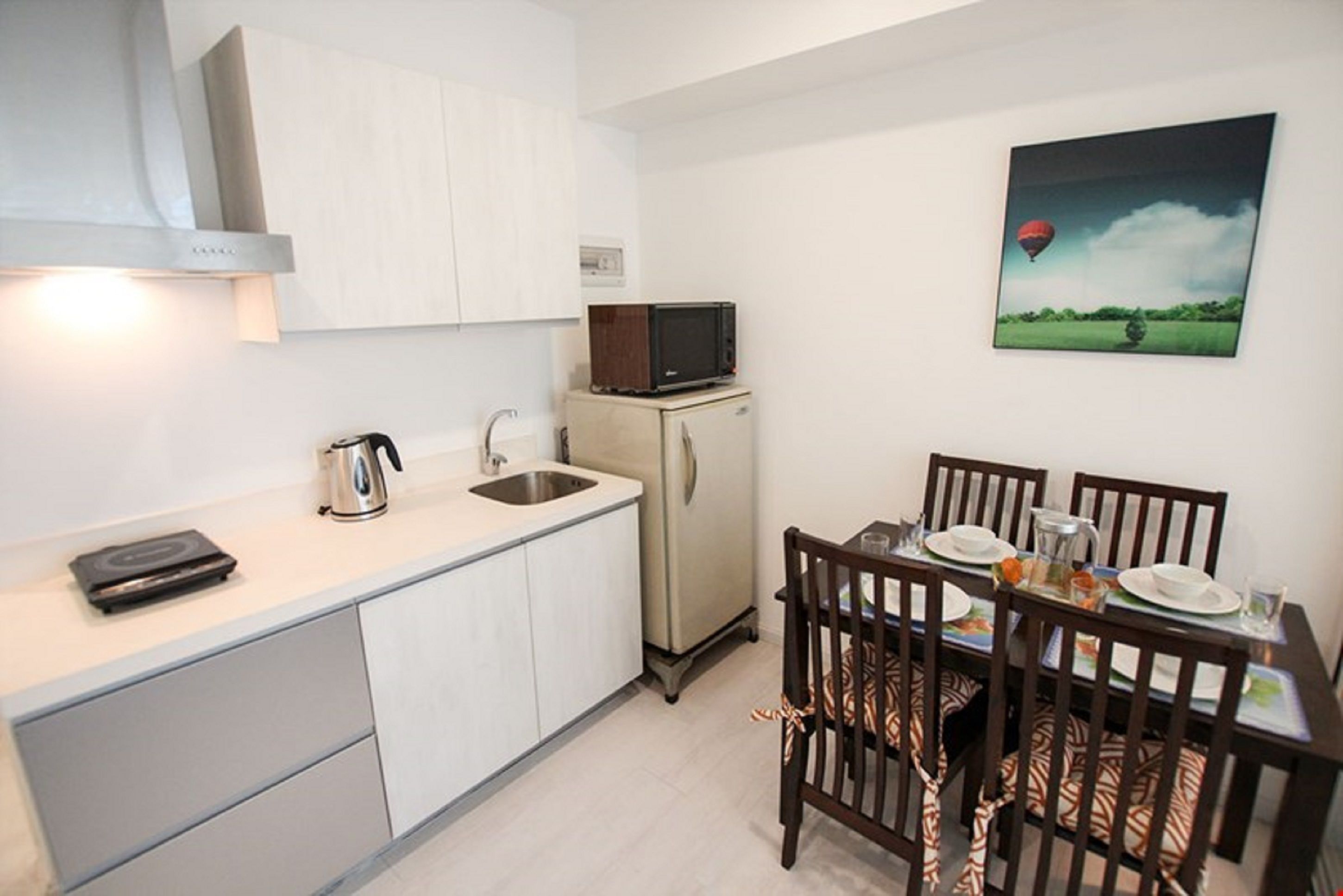 Cocina compacta con microondas, hervidor y un refrigerador pequeño. Hay una mesa de comedor con sillas y un cuadro de un globo aerostático en la pared.