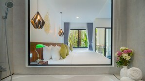 Un baño moderno con un espejo grande, una bañera llena de pétalos de rosa y vistas a un dormitorio con cama, almohadas y ventana.