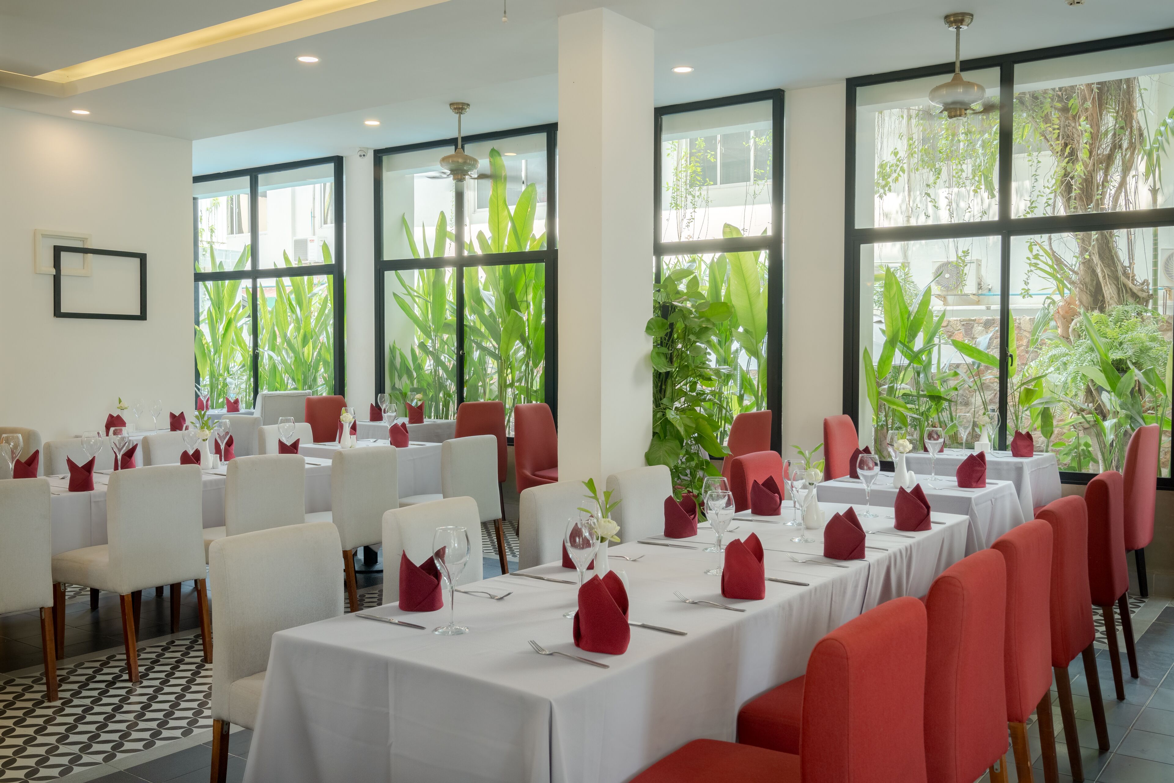 Un restaurante con mesas blancas y sillas rojas, ventanas amplias y plantas de interior.