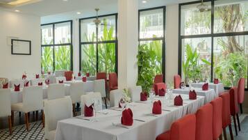Un restaurante con mesas blancas y sillas rojas, ventanas amplias y plantas de interior.