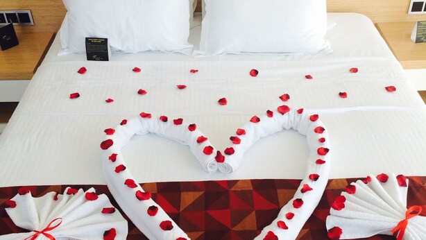 Una cama bien hecha con sábanas blancas, un arreglo de pétalos de rosa en forma de corazón y dos toallas adornadas con pétalos de rosa.