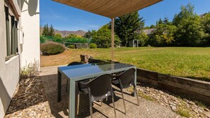 Une terrasse extérieure avec une table et des chaises, un auvent pare-soleil et une vue sur une zone herbeuse, avec des arbres et des montagnes en arrière-plan.