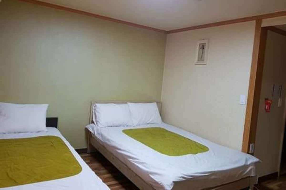 Um quarto pequeno com duas camas individuais, cada uma com colchão branco e manta verde. O quarto possui piso de madeira e teto branco com claraboia.