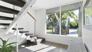 A modern staircase with a glass railing leading to a balcony, a wicker bench, and a view of a garden and street.