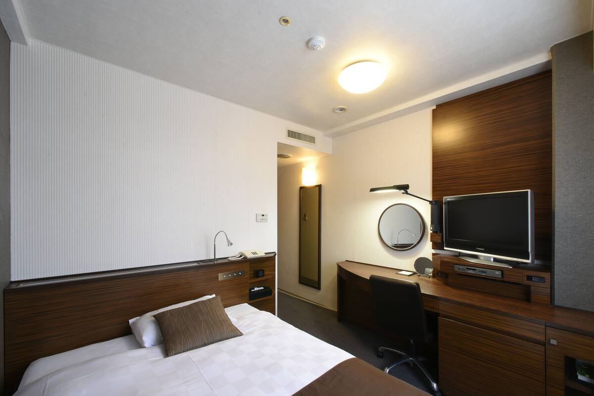 A hotel room with a bed, a desk with a TV, a mirror, and a chair.