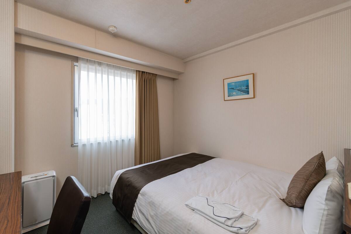 A hotel room with a bed, a desk, a chair, a radiator, a window with curtains, and a picture on the wall.
