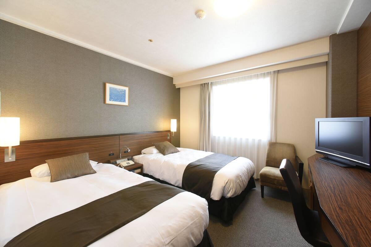 A hotel room with two beds, a television, a chair, and a window with curtains.