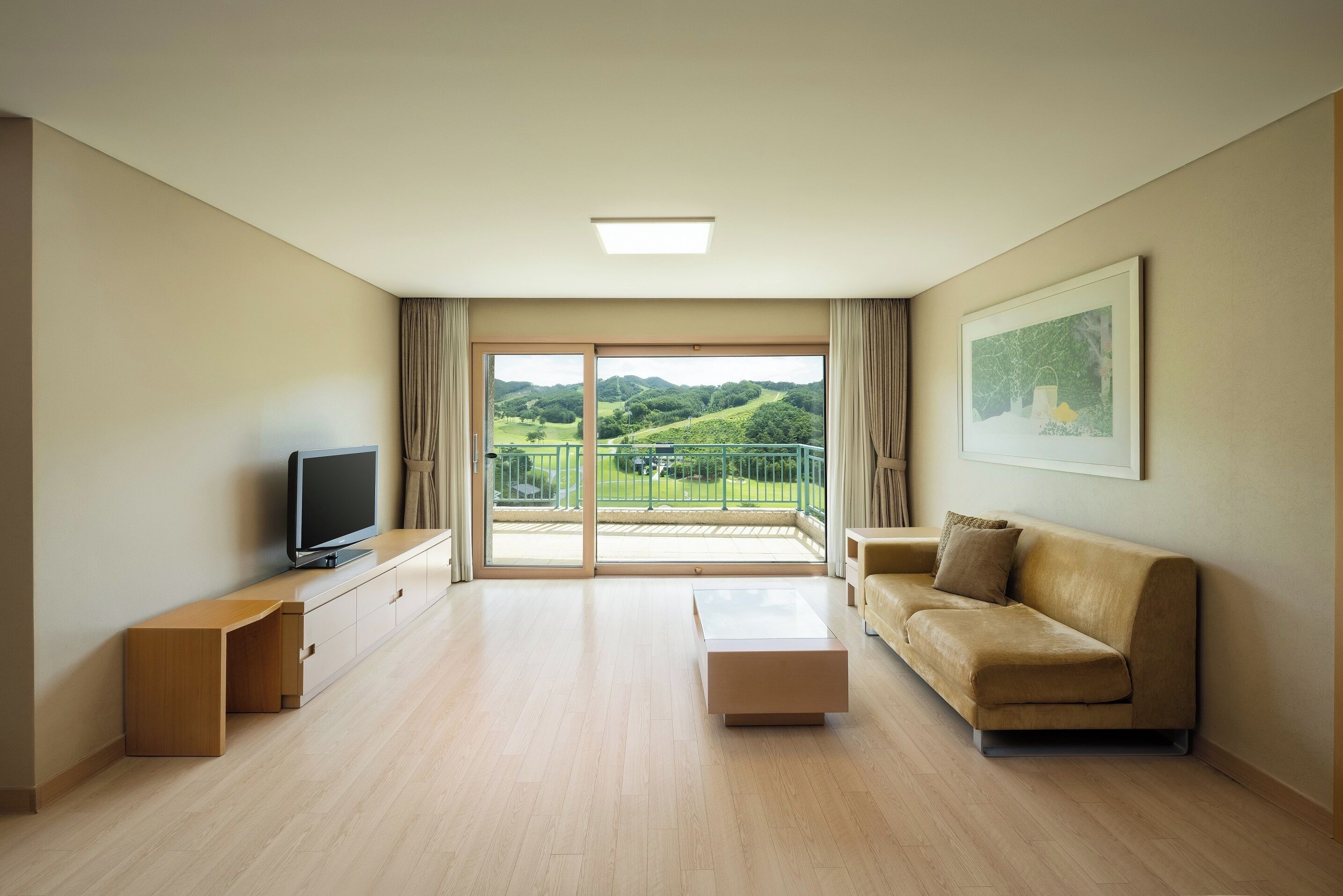 A modern living room with a flat-screen TV, a sofa, a coffee table, and a sliding glass door leading to a balcony with a view of greenery.