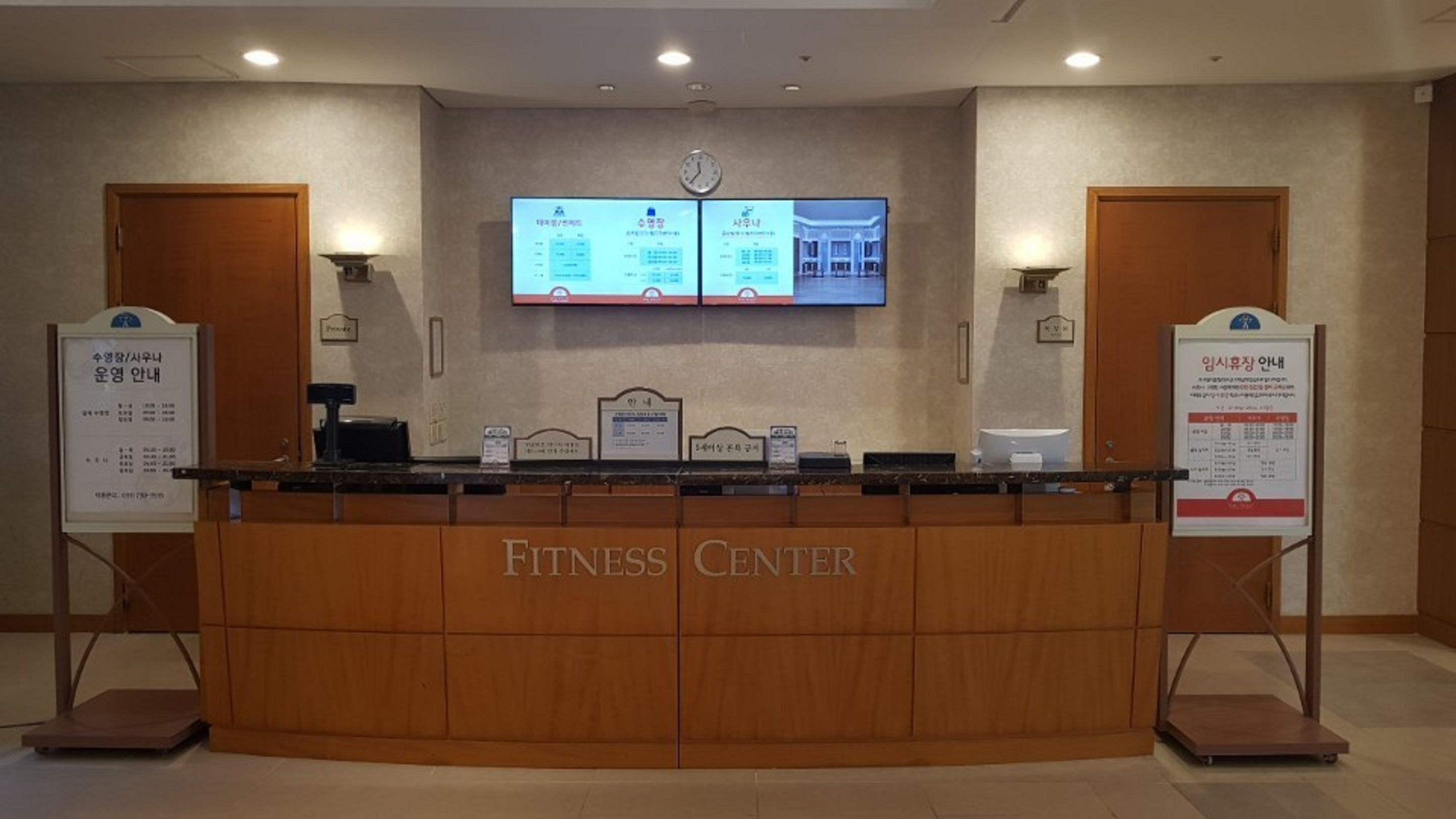 A reception desk with a digital screen displaying information, wooden panels, and a clock on the wall.
