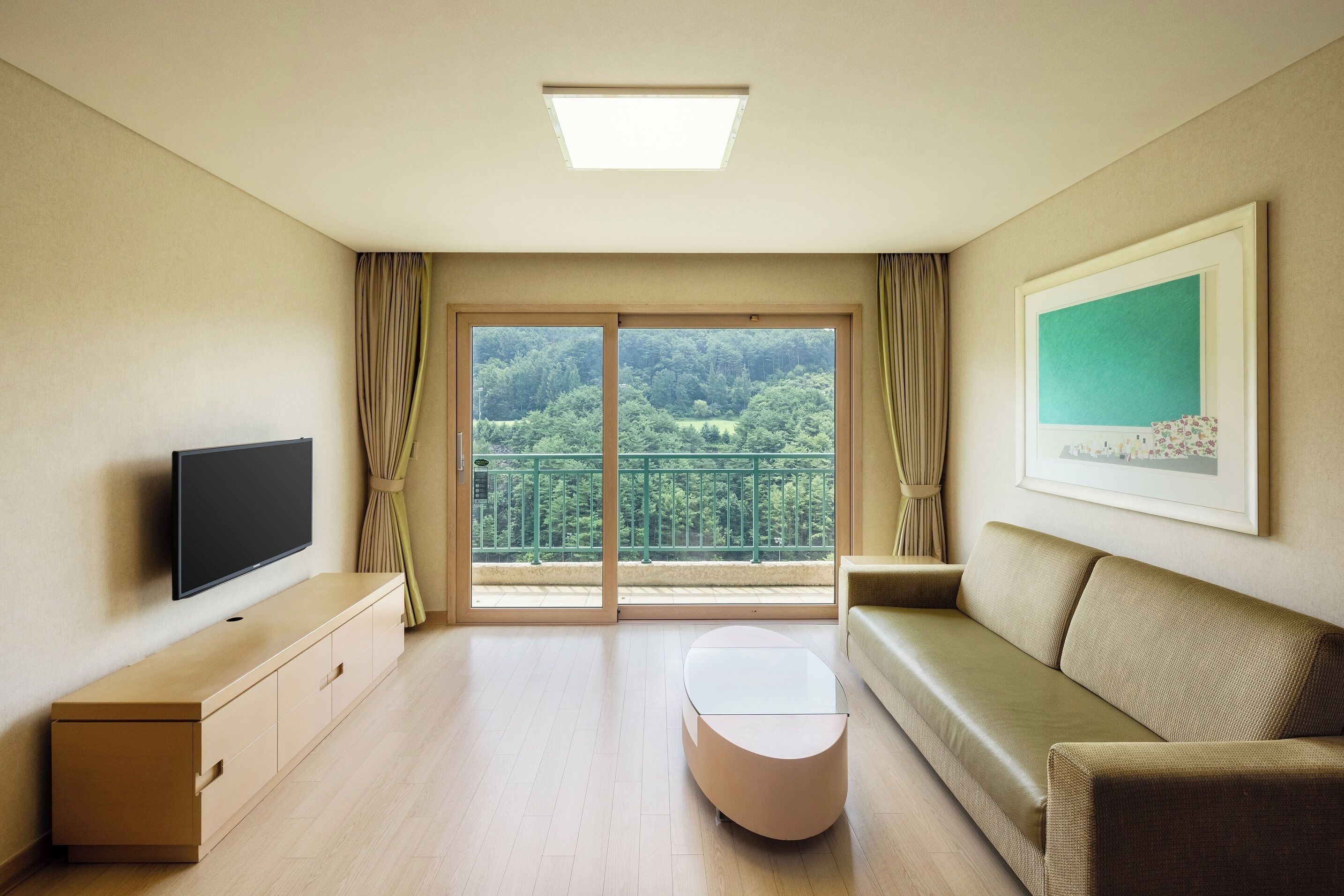 Una sala de estar moderna con un televisor de pantalla plana, un sofá, una mesa de centro y un amplio ventanal corredero que da a un balcón con vistas a un bosque.