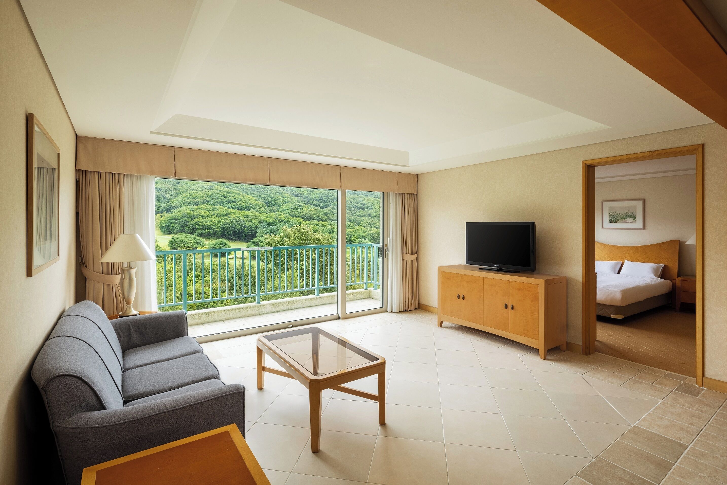 A modern hotel room with a sofa, a glass coffee table, a TV on a wooden cabinet, and a balcony with a view of greenery.