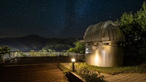 A telescope dome with a wooden deck and railing, surrounded by greenery and mountains under a starry night sky. - Oak Valley Valley Village (Wonju)