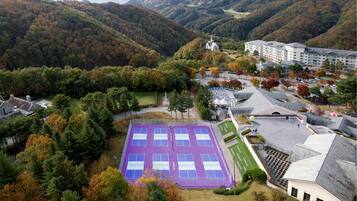 A tennis court surrounded by a forested area with a church and residential buildings in the background.