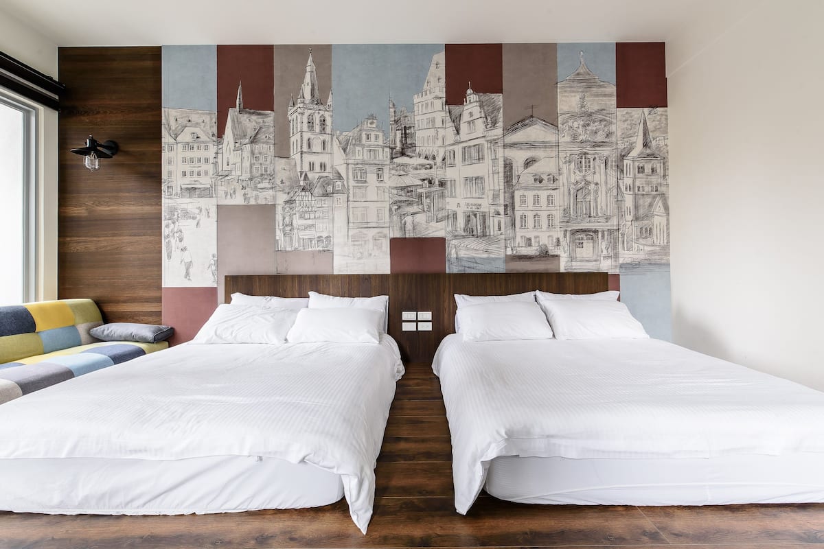Quarto de hotel com duas camas, uma grande obra de arte urbana na parede, cabeceira de madeira e um sofá colorido.