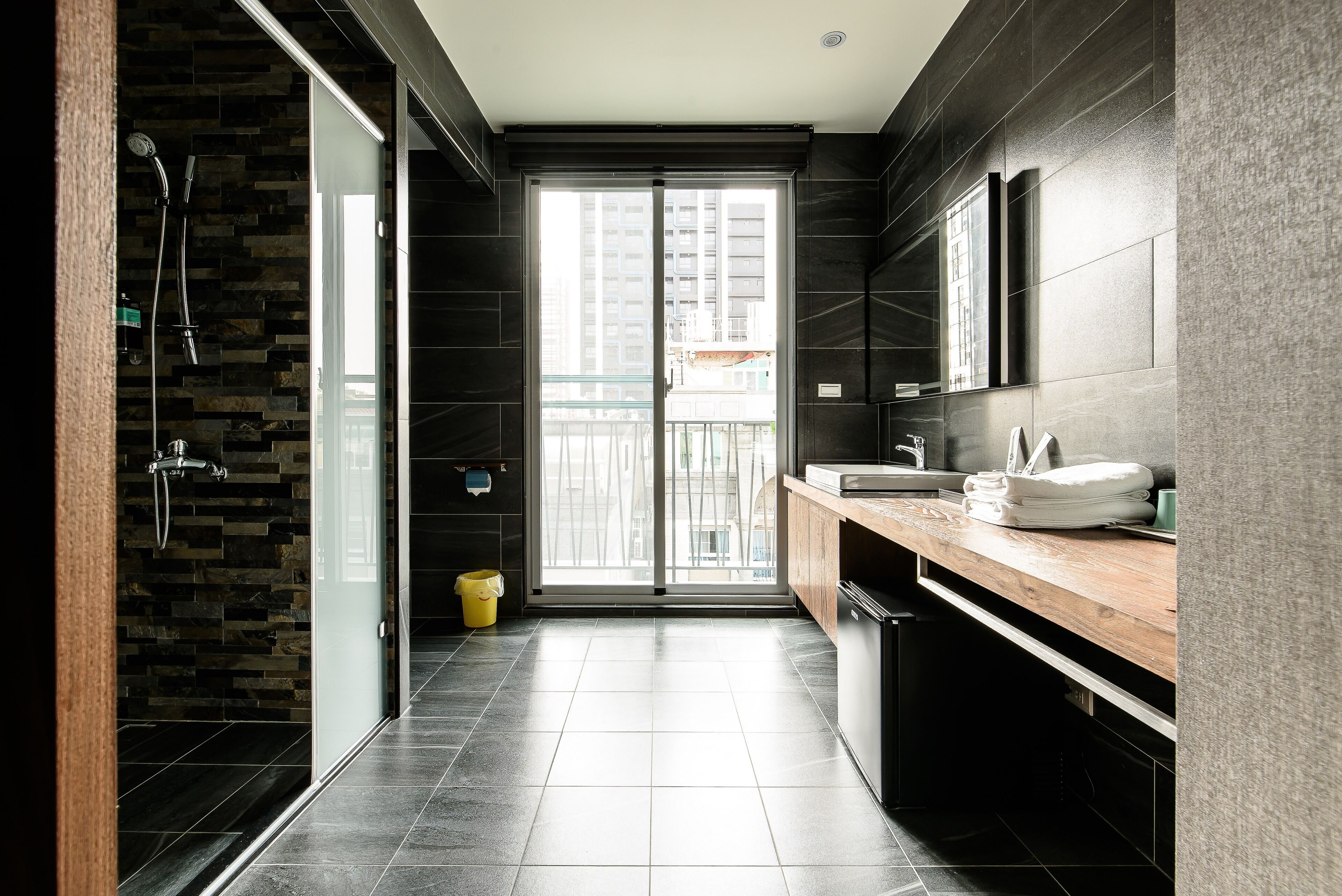A modern bathroom with a glass door leading to a balcony, a wooden vanity, and a shower area with a stone wall.