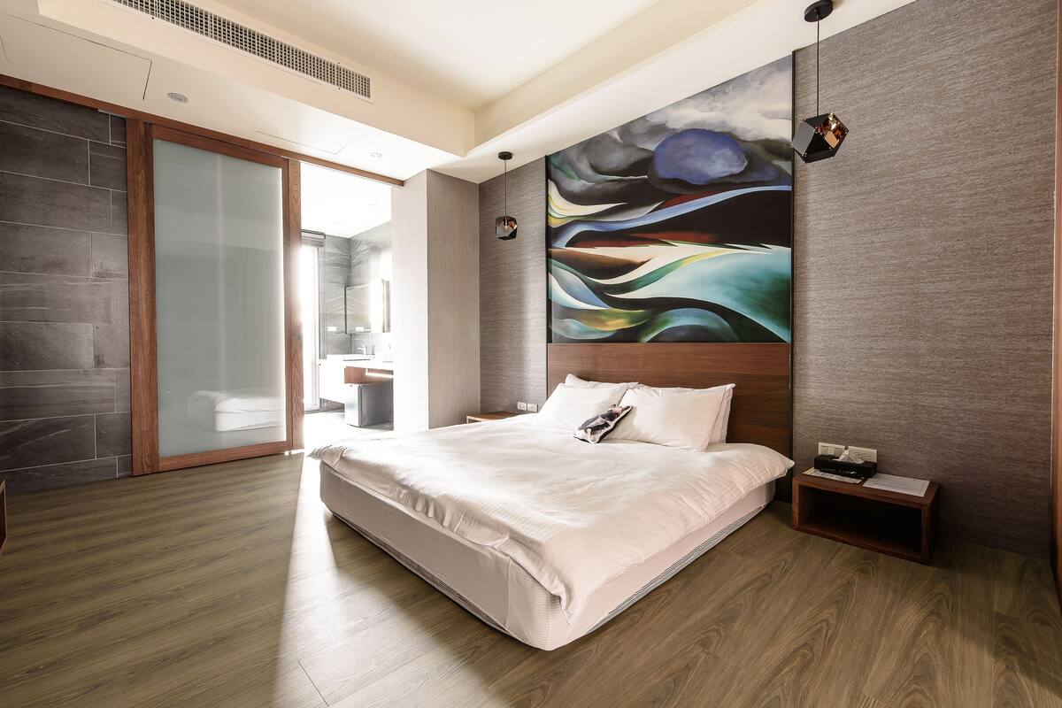 Quarto moderno com uma cama grande, mesa de cabeceira, um quadro e porta deslizante para um banheiro.