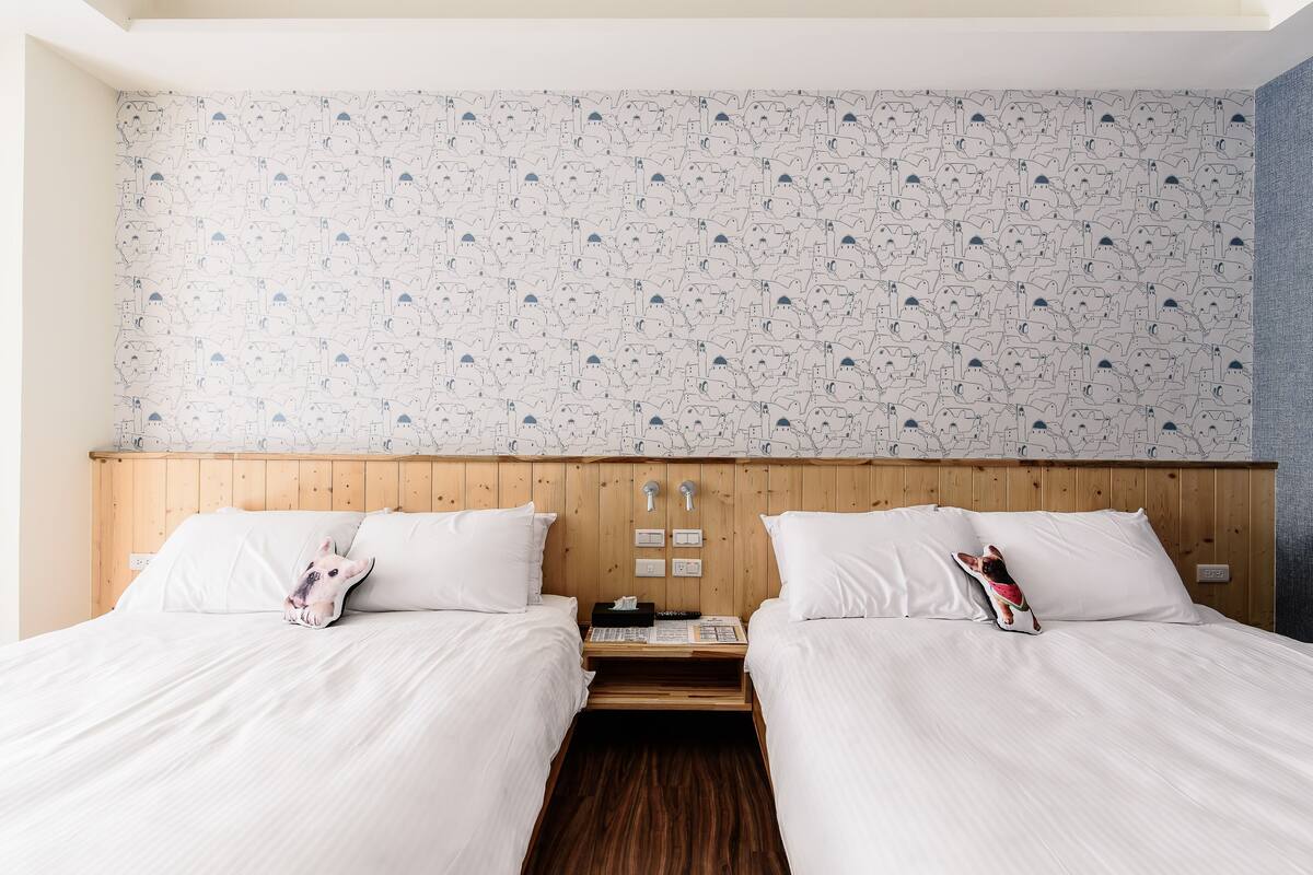 Quarto de hotel com duas camas, cabeceira de madeira, roupa de cama branca e papel de parede estampado.