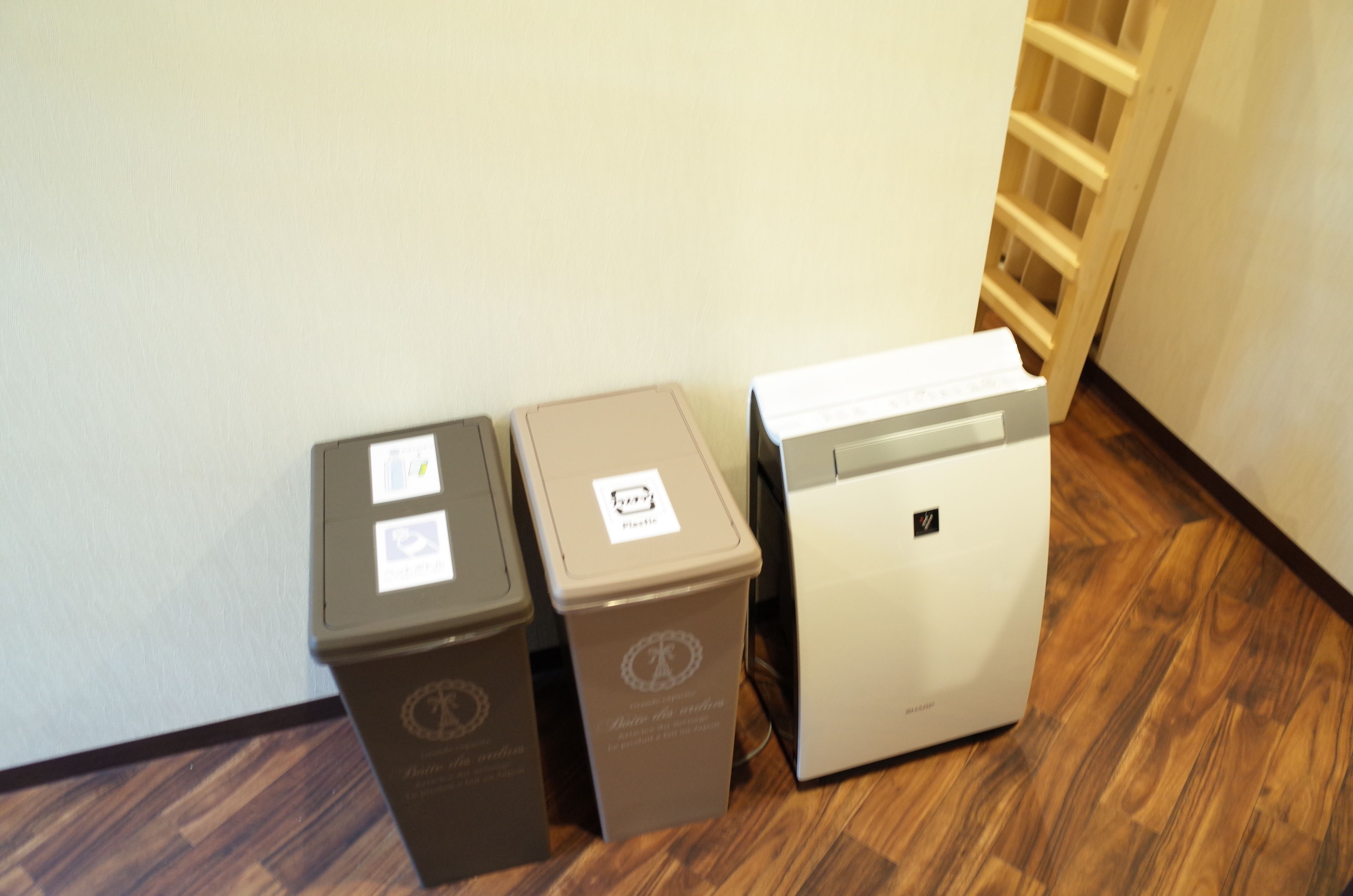 Three trash bins and a white appliance, likely a coffee machine, in a hallway with wooden flooring and a white wall.