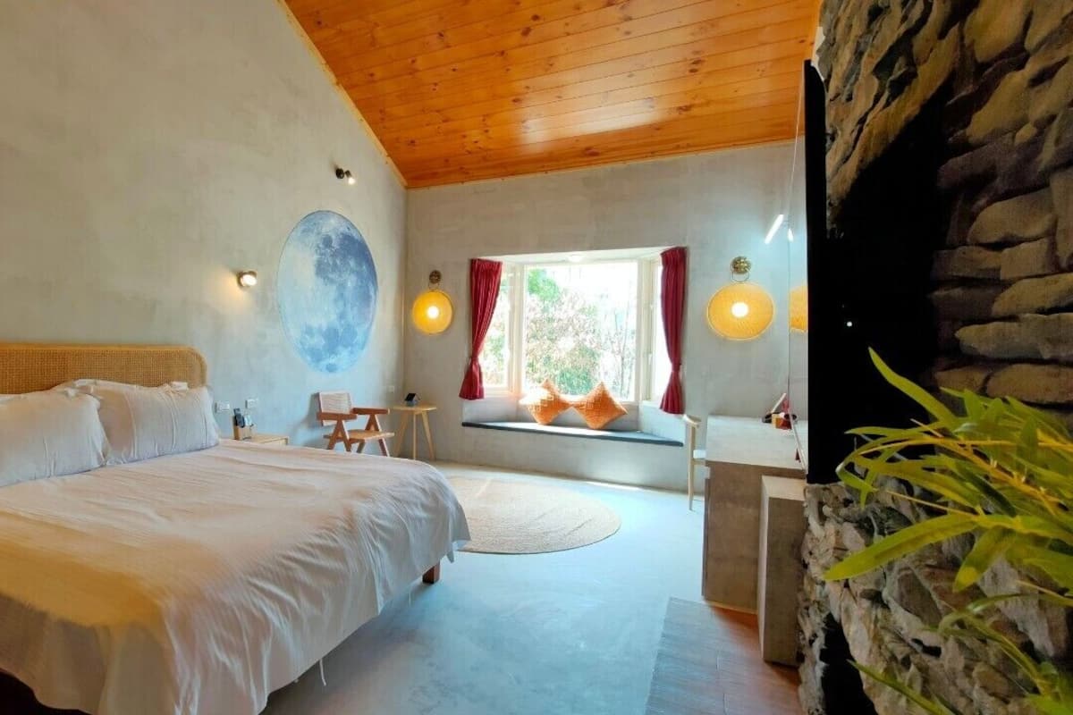 A bedroom with a large bed, a small table, a window with curtains, and a stone wall.