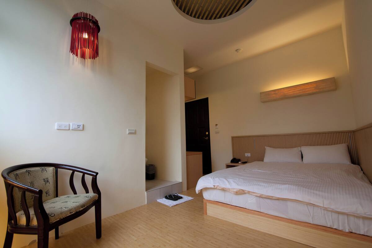 A hotel room with a wooden bed, a chair, a desk, and a red hanging light fixture.