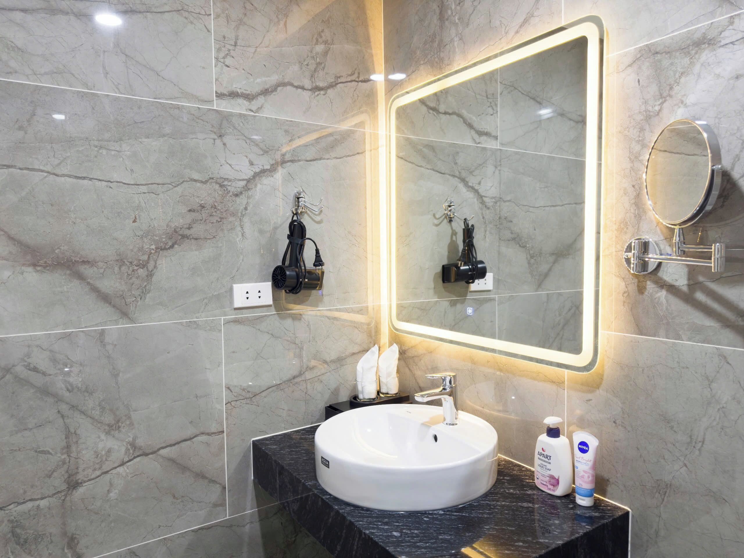 Un baño moderno con piso de mármol, un lavamanos blanco y un espejo con marco iluminado.