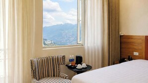 Une chambre d’hôtel avec un lit, un fauteuil à rayures, une petite table avec une machine à café et des tasses, et une fenêtre donnant sur la montagne.