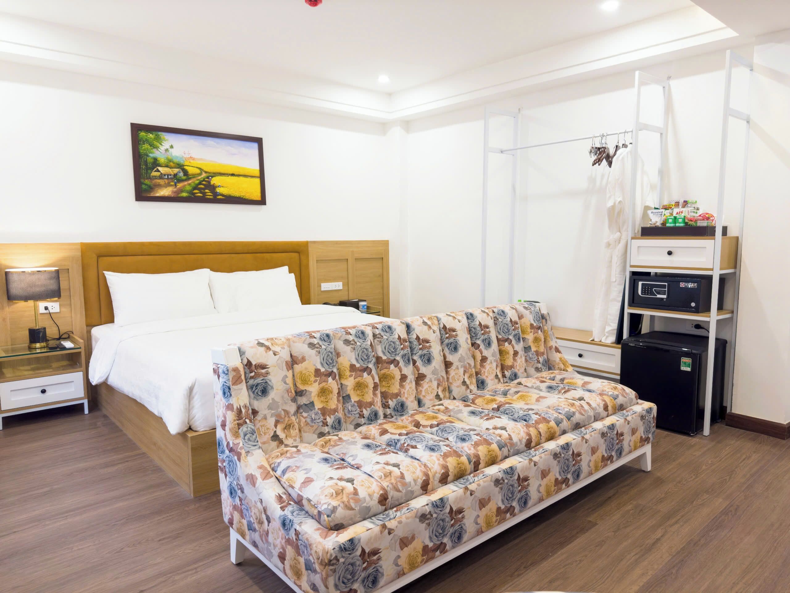 A hotel room with a bed, a sofa, a nightstand, a lamp, a painting, a wardrobe, and a refrigerator.