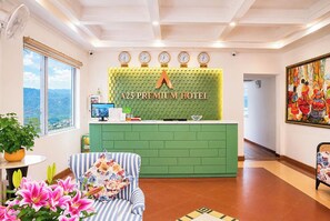 Zona de recepción de hotel con un mostrador verde, relojes mostrando diferentes husos horarios, un monitor de computadora y un amplio ventanal con vista al paisaje.