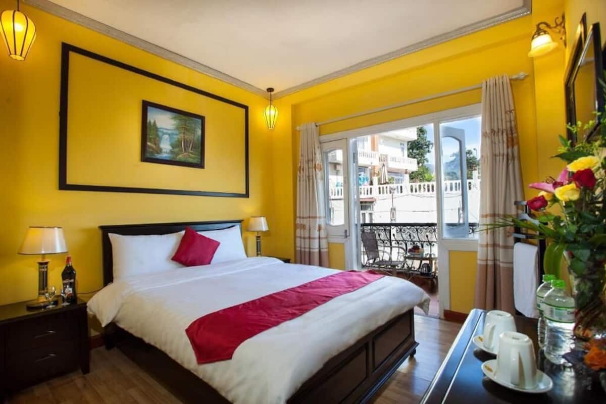 Una habitación de hotel con una cama, una mesita de noche, una mesa con un florero y un balcón con vistas.