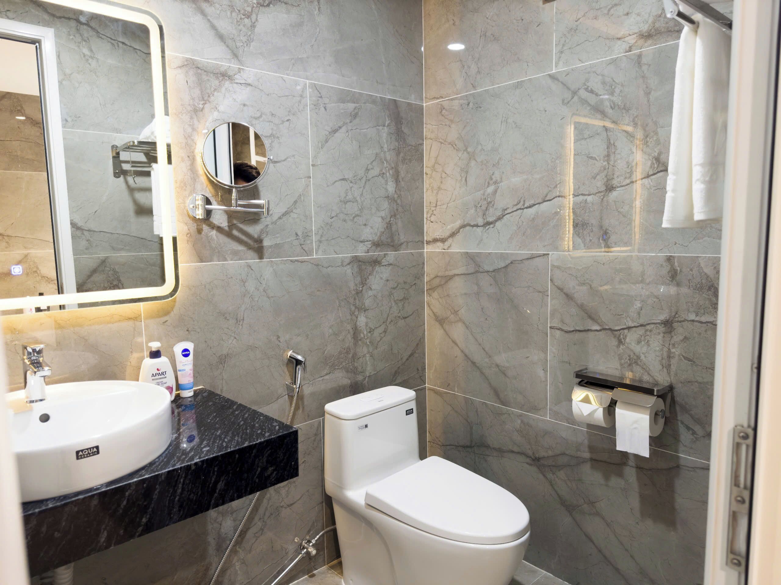 A modern bathroom with a marble backsplash, a round mirror, a white sink, and a toilet.