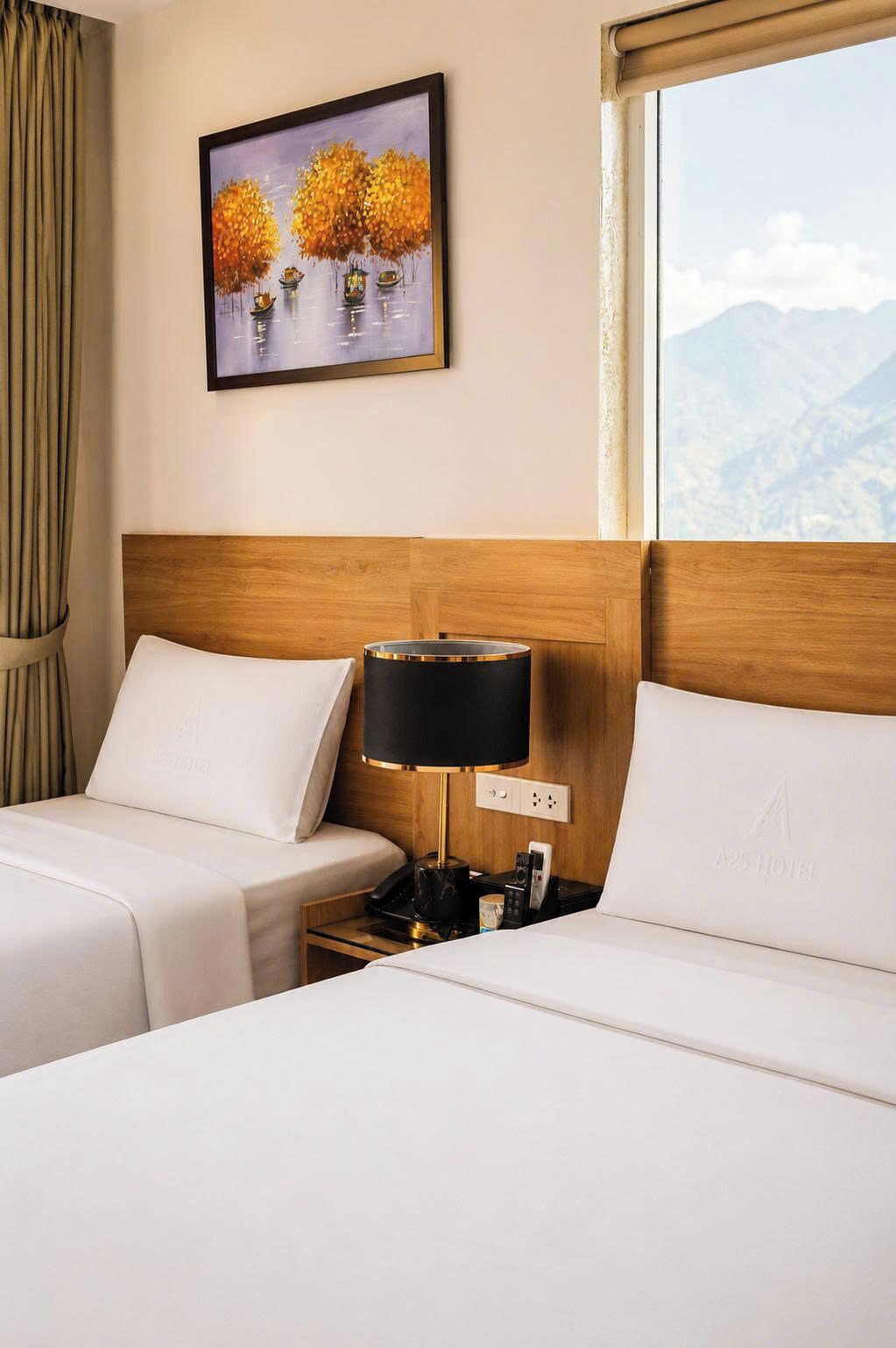 A hotel room with two beds, a nightstand with a lamp, a painting of trees, and a view of mountains.