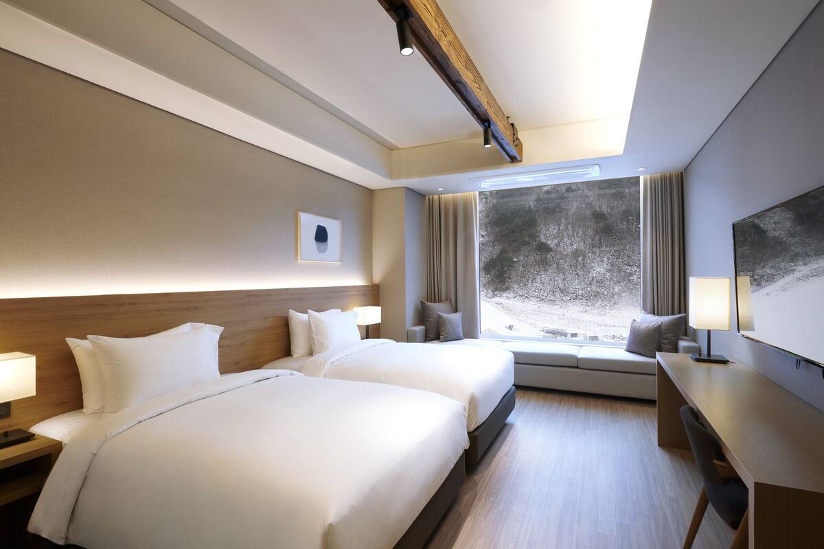 A hotel room with two beds, a desk, a chair, a window with a view of trees, and a mounted artwork on the wall.
