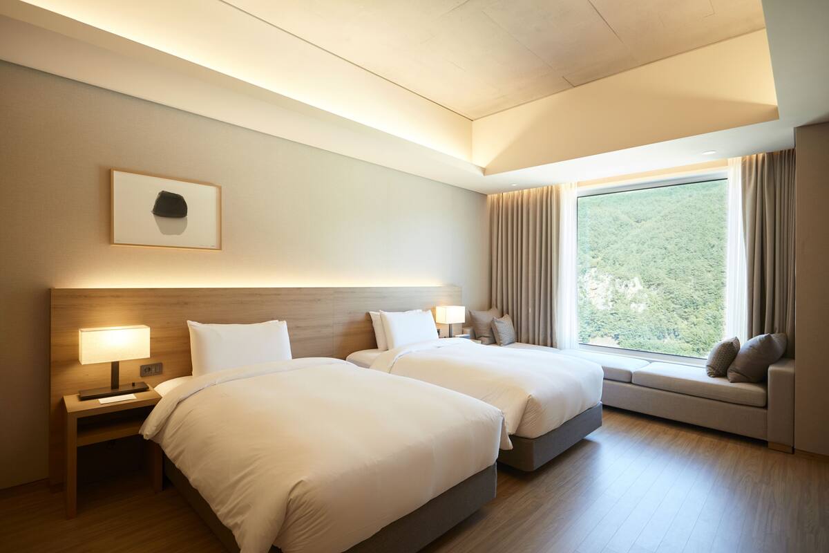 A hotel room with two beds, a nightstand with a lamp, a window with curtains, and a view of greenery outside.