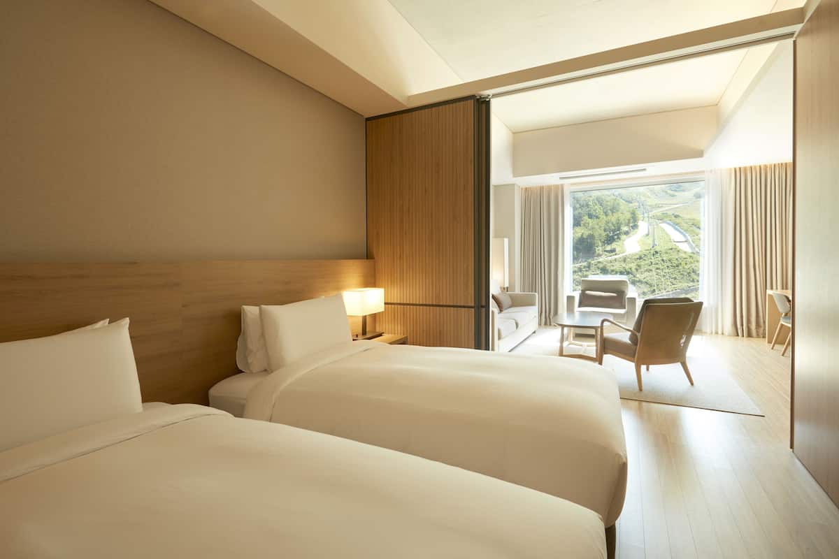 A hotel room with two beds, a wooden headboard, a nightstand with a lamp, and a balcony with a view of greenery and a road.