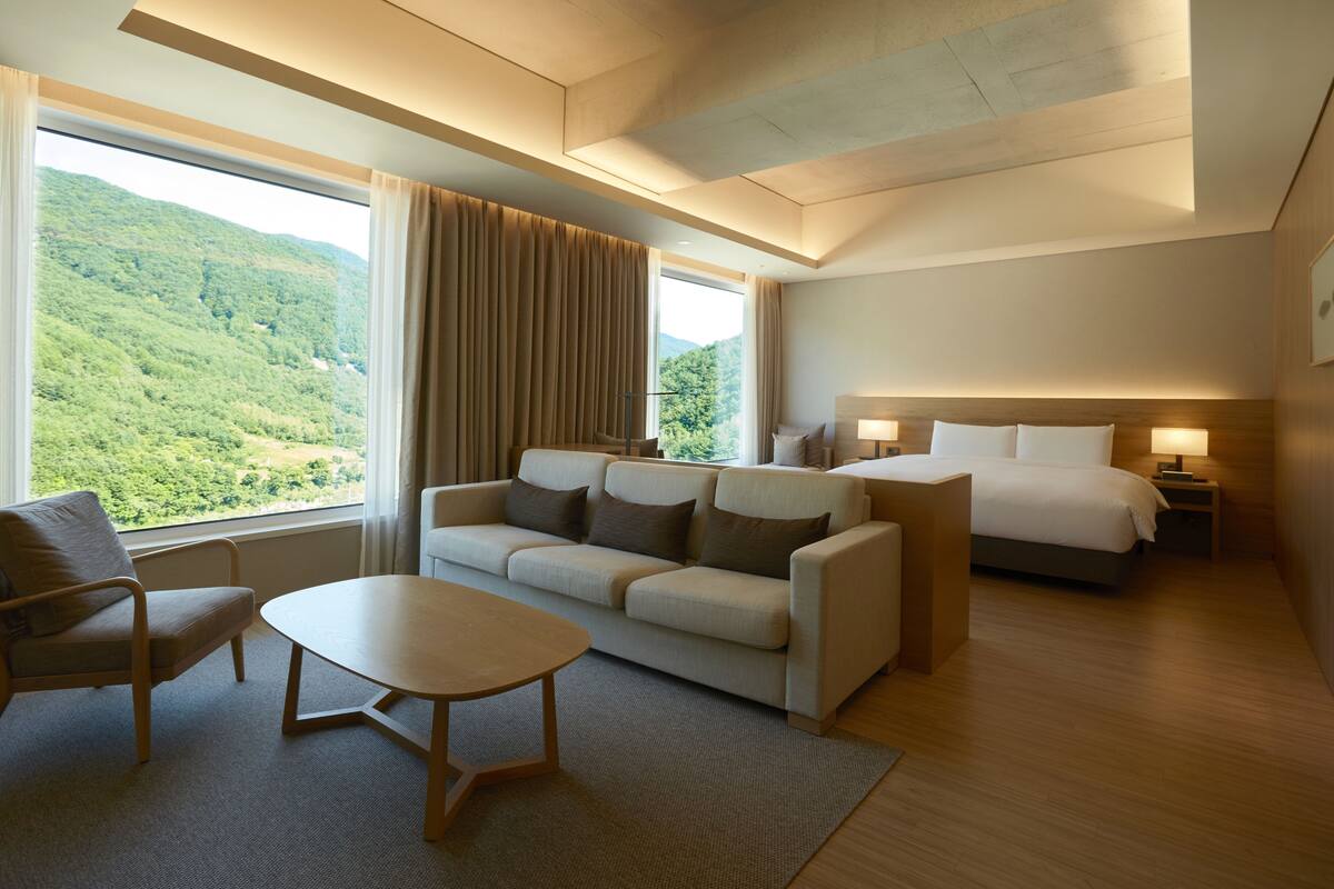 A hotel room with a large window offering a view of a forested hillside, a sofa, a bed, a bedside table, and a nightstand with a lamp.