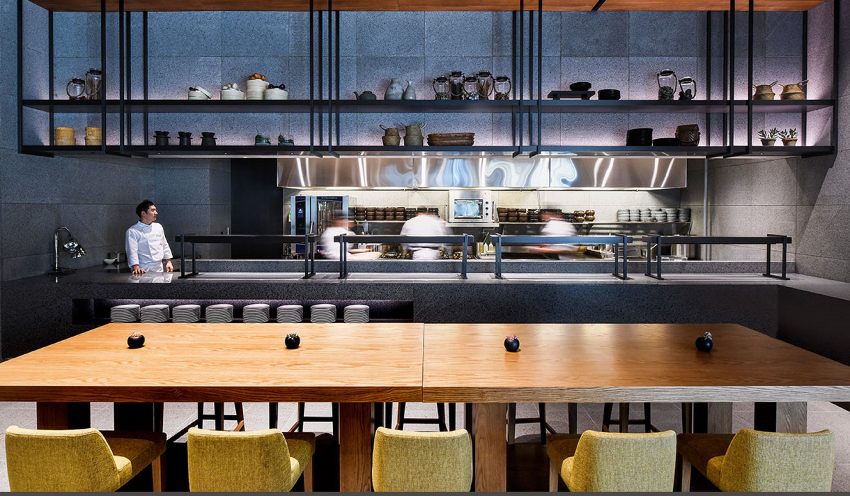 Een modern restaurant met een lange houten tafel en gele stoelen, een kok op de achtergrond en planken met keukengerei.