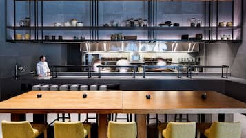 Un restaurante moderno con una larga mesa de madera y sillas amarillas, un chef al fondo y estanterías con utensilios de cocina.
