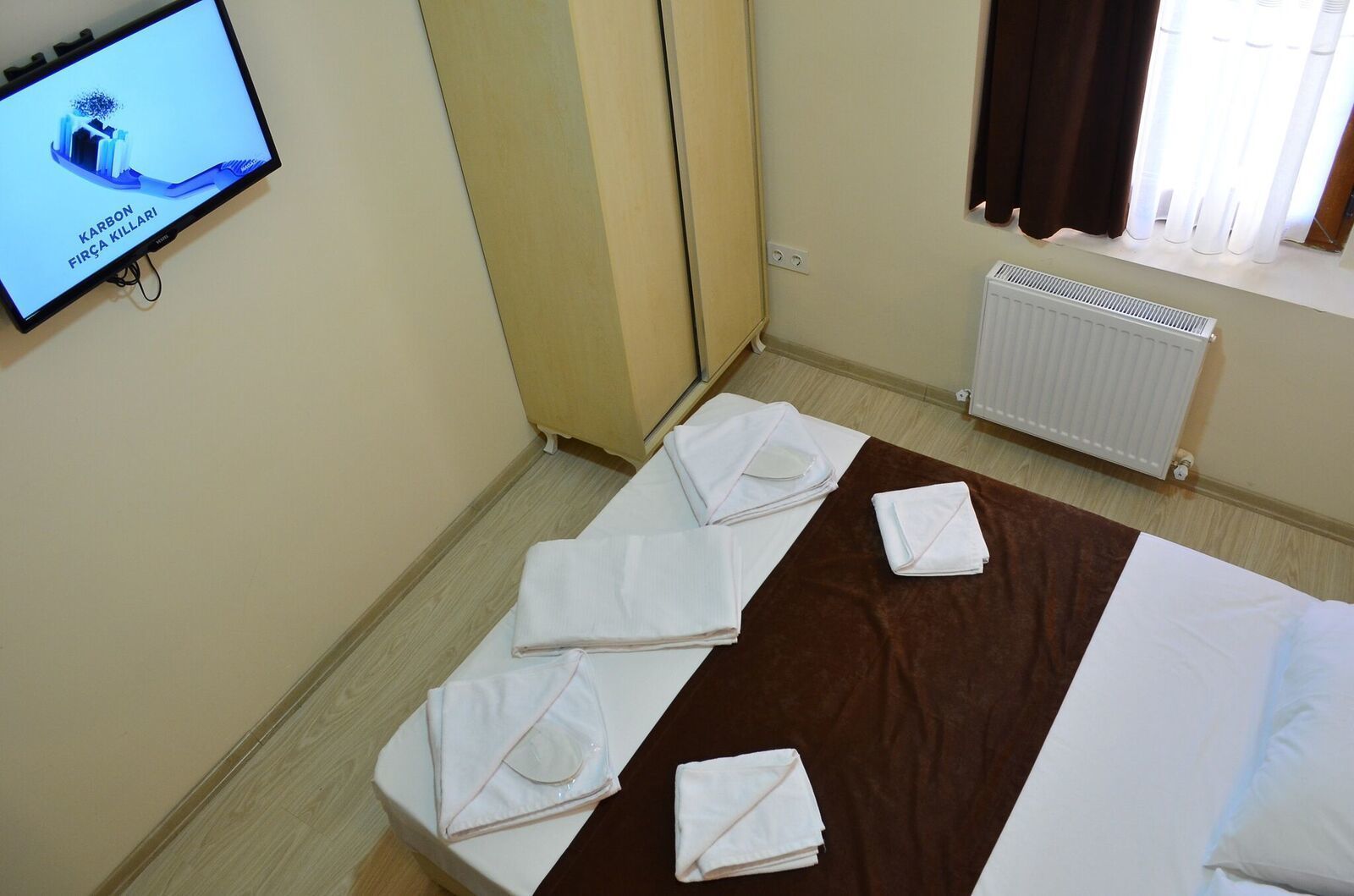 Habitación de hotel con cama, televisor mostrando 'KARBON FIRÇA KILLARI', radiador y una alfombra marrón.