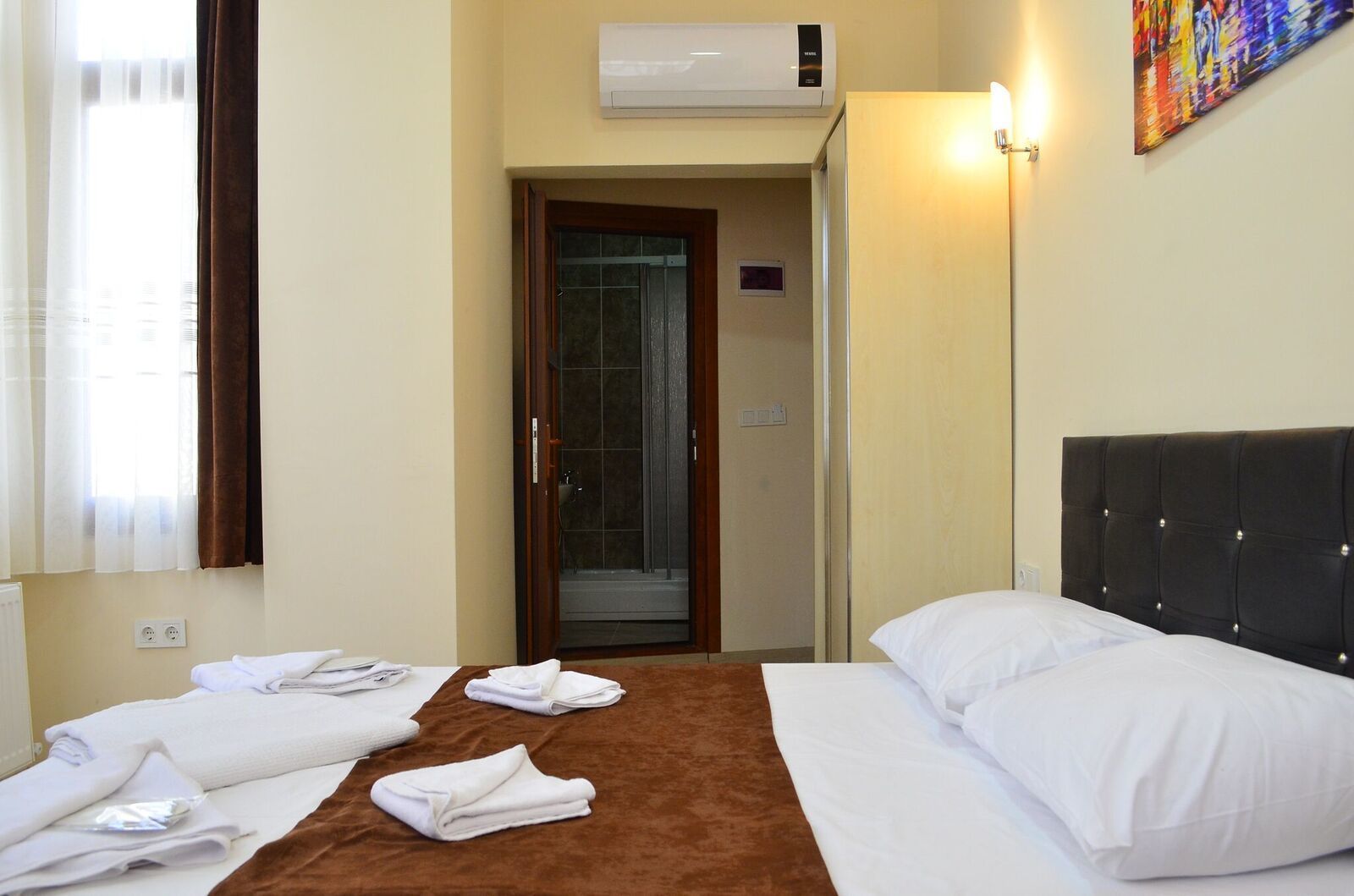 A hotel room with a bed, two towels, a door, an air conditioning unit, and a painting on the wall.