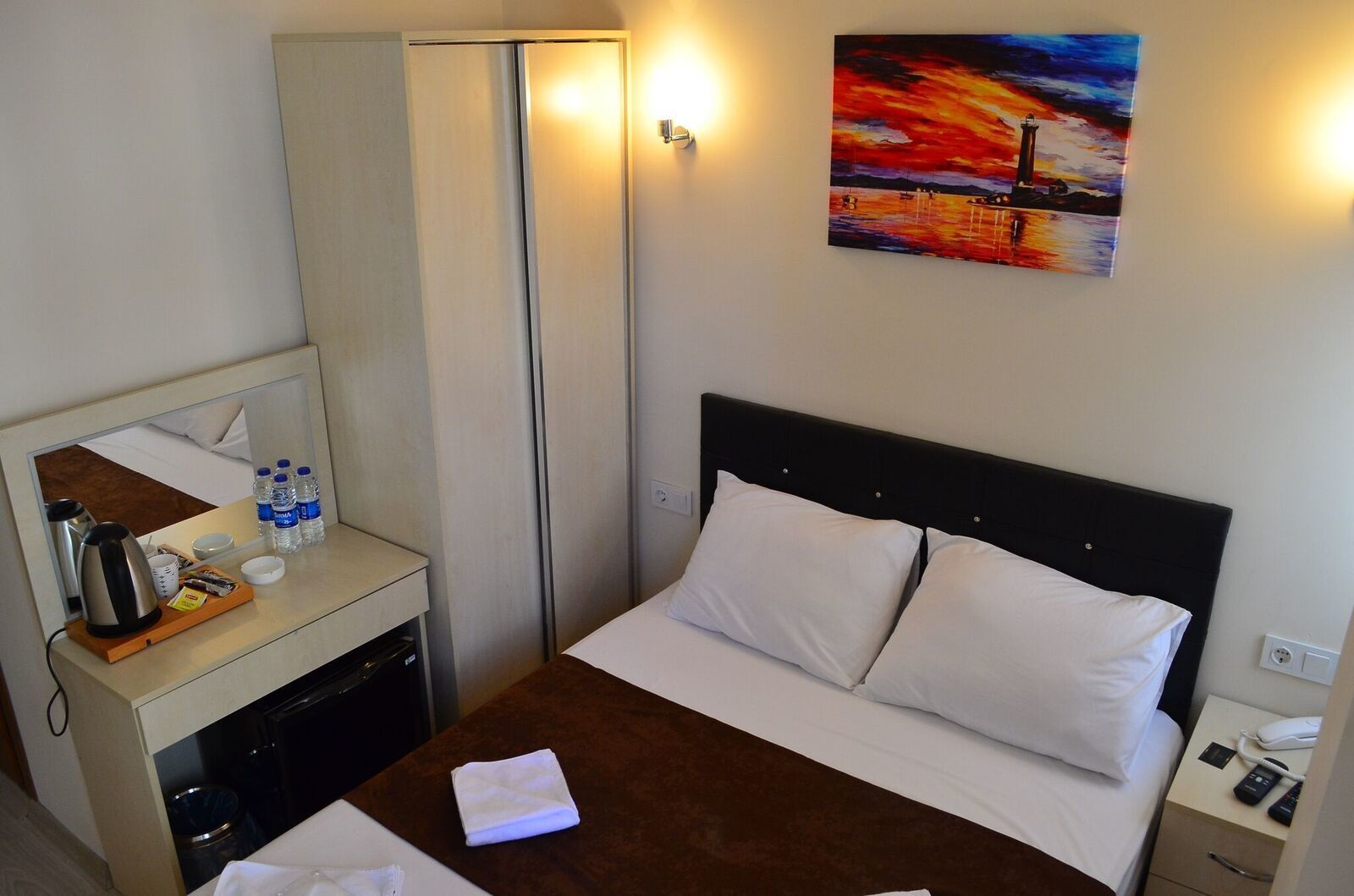 A hotel room with a bed, a bedside table with a kettle, cups, and bottled water, a mirror, a painting of a sunset, and a nightstand with a phone.