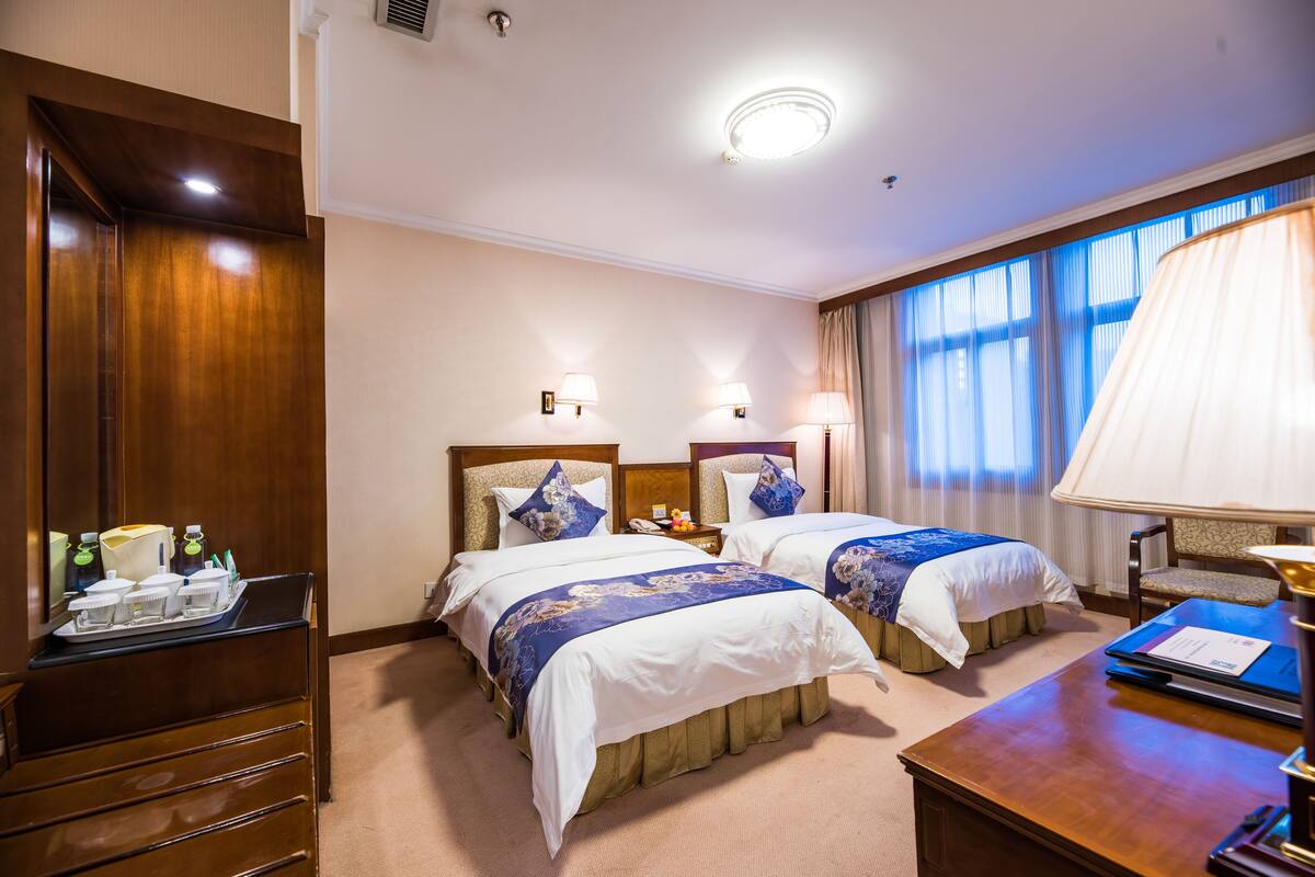 Une chambre d’hôtel avec deux lits, un bureau, une chaise, une table de chevet et une fenêtre avec des rideaux.