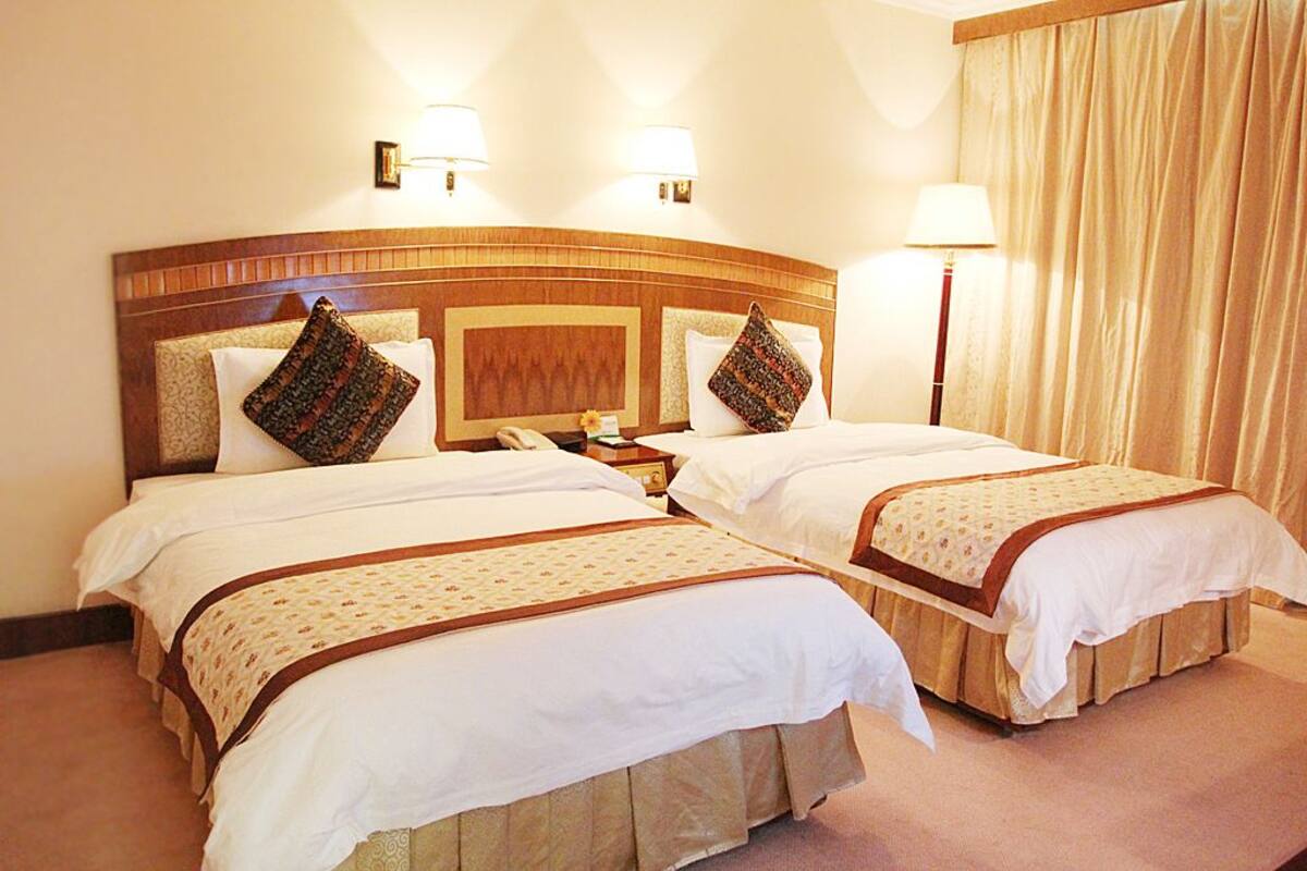 Une chambre d’hôtel avec deux lits, chacun doté d’une tête de lit en bois, de draps blancs et de coussins décoratifs à motifs. On y trouve des lampes de chevet et une table de chevet avec une lampe.