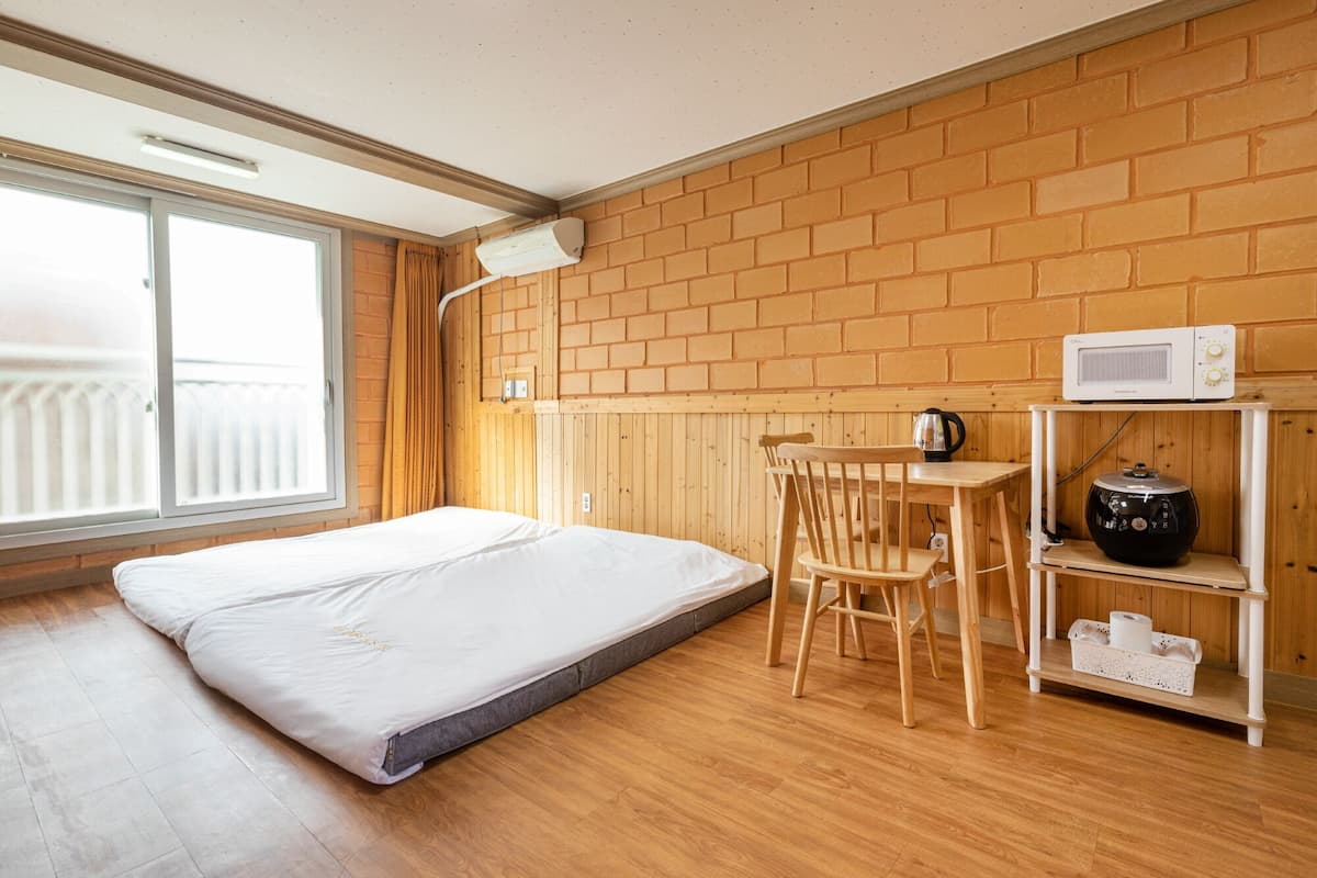 A room with a wooden floor, a bed, a table with chairs, a microwave, and a shelf with a kettle and other items.