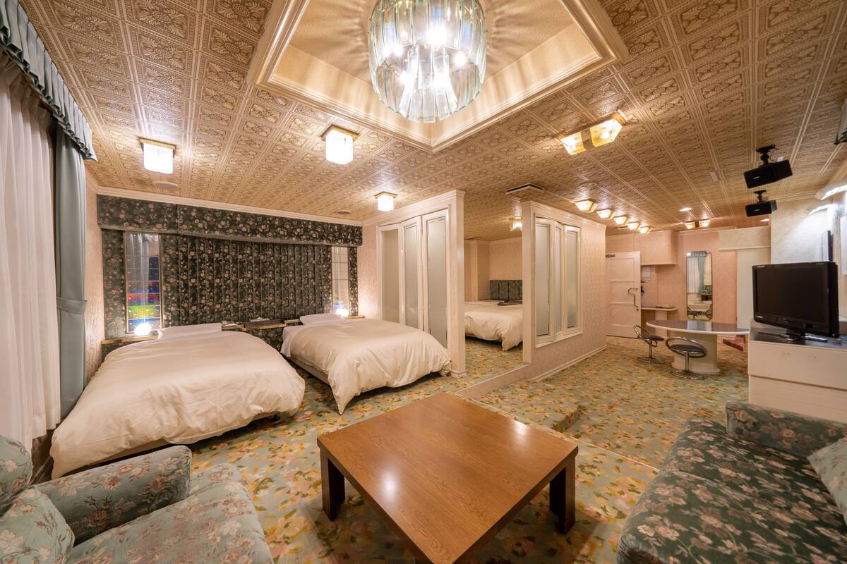 Ein Hotelzimmer mit zwei Betten, einem Holztisch, einer Couch, einem Spiegel, einem Fernseher und einer Decke mit aufwendigen Mustern.