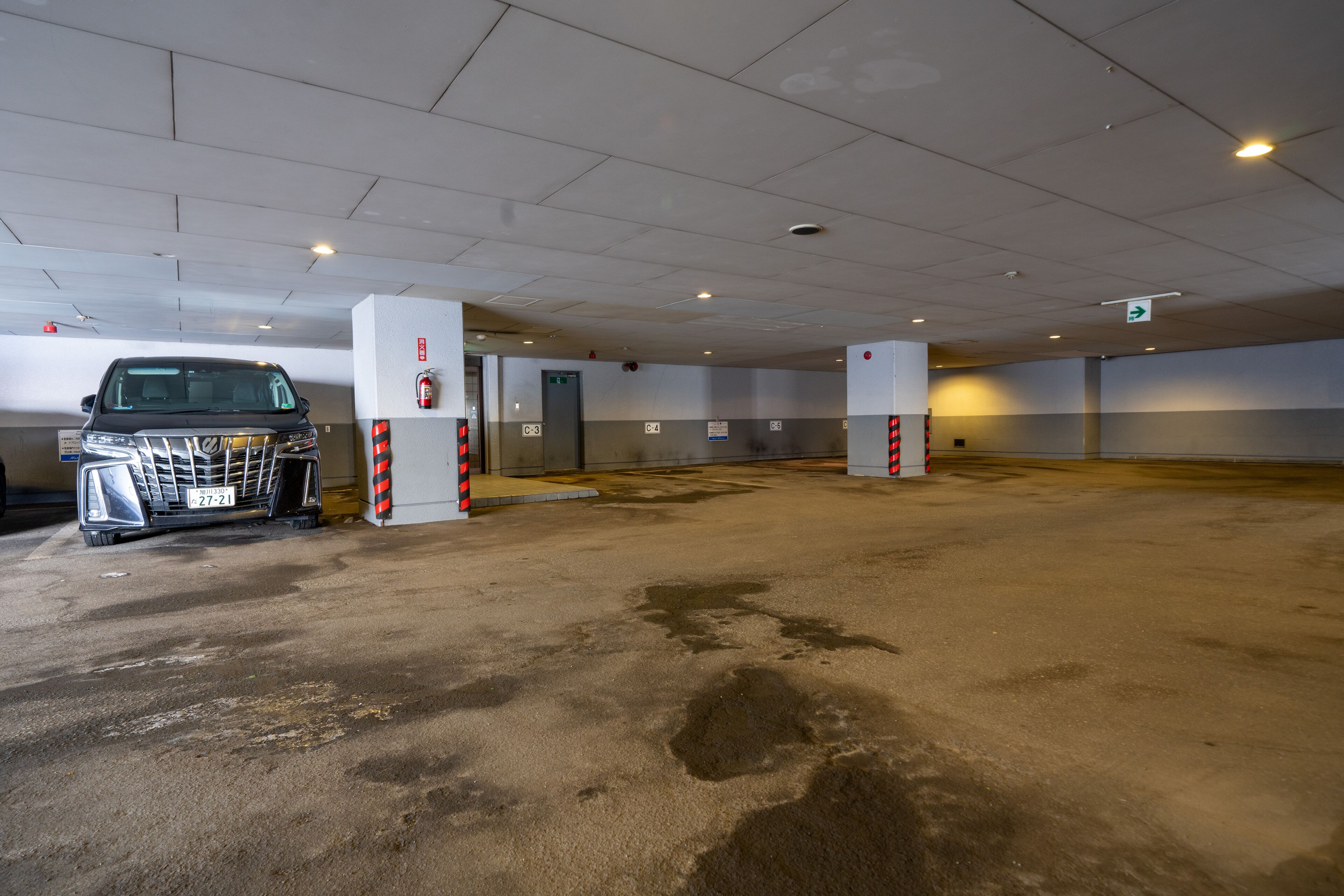 A parking garage with a car parked near a pillar, emergency exit signs, and a fire extinguisher on the wall.