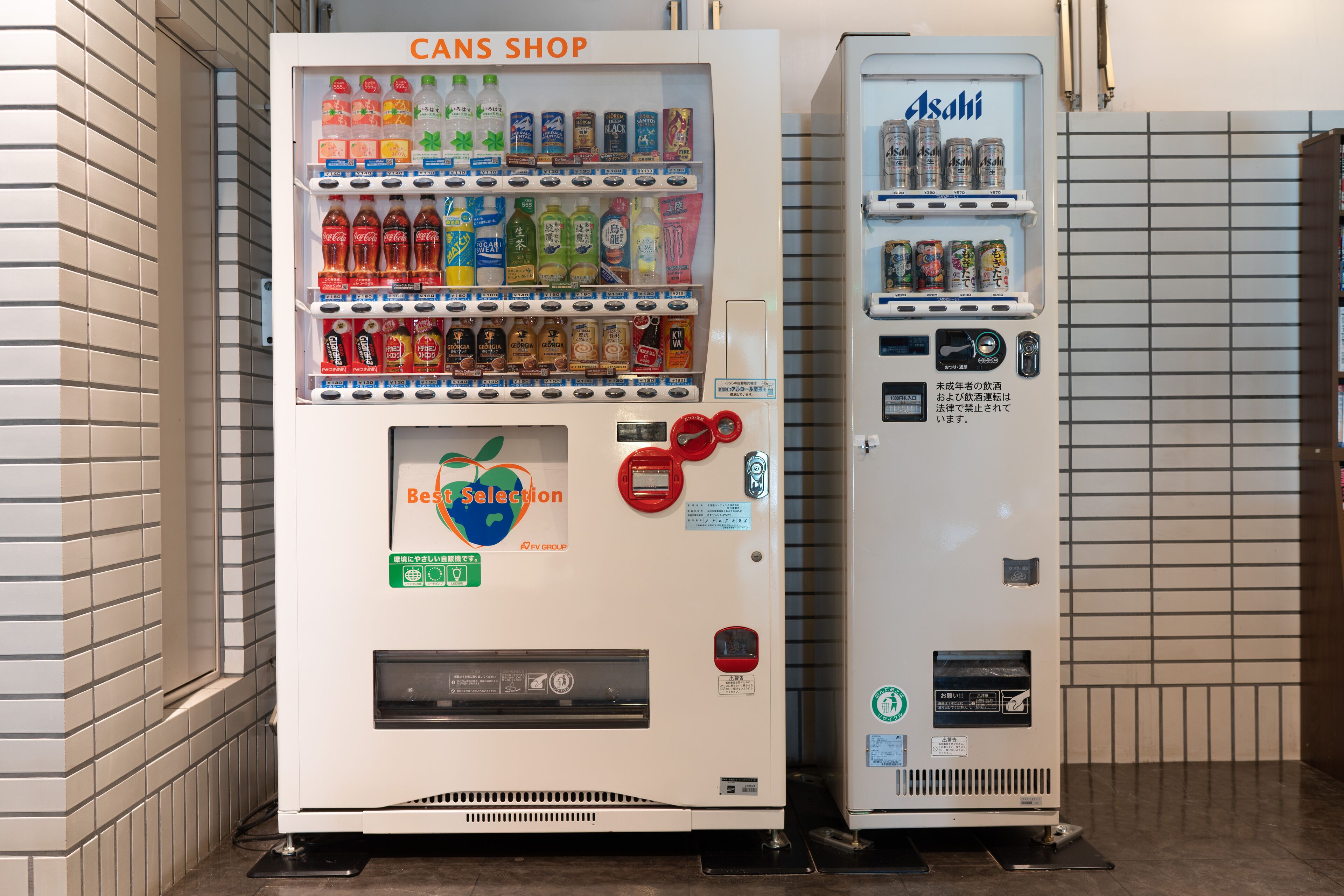 Deux distributeurs automatiques, l’un portant l’inscription « Cans Shop » et l’autre « Asahi », proposent une variété de boissons et d’en-cas.