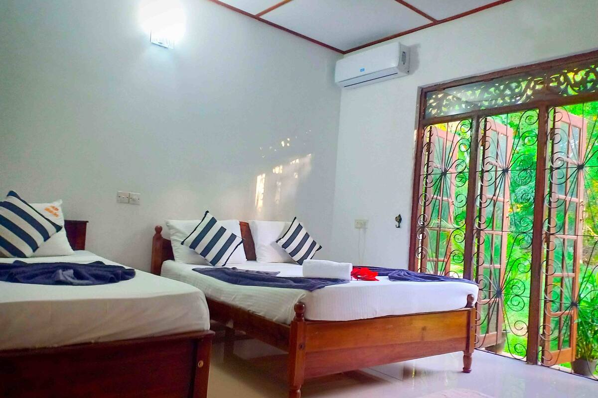 A room with two wooden beds, white walls, an air conditioning unit, and a view through a metal gate.