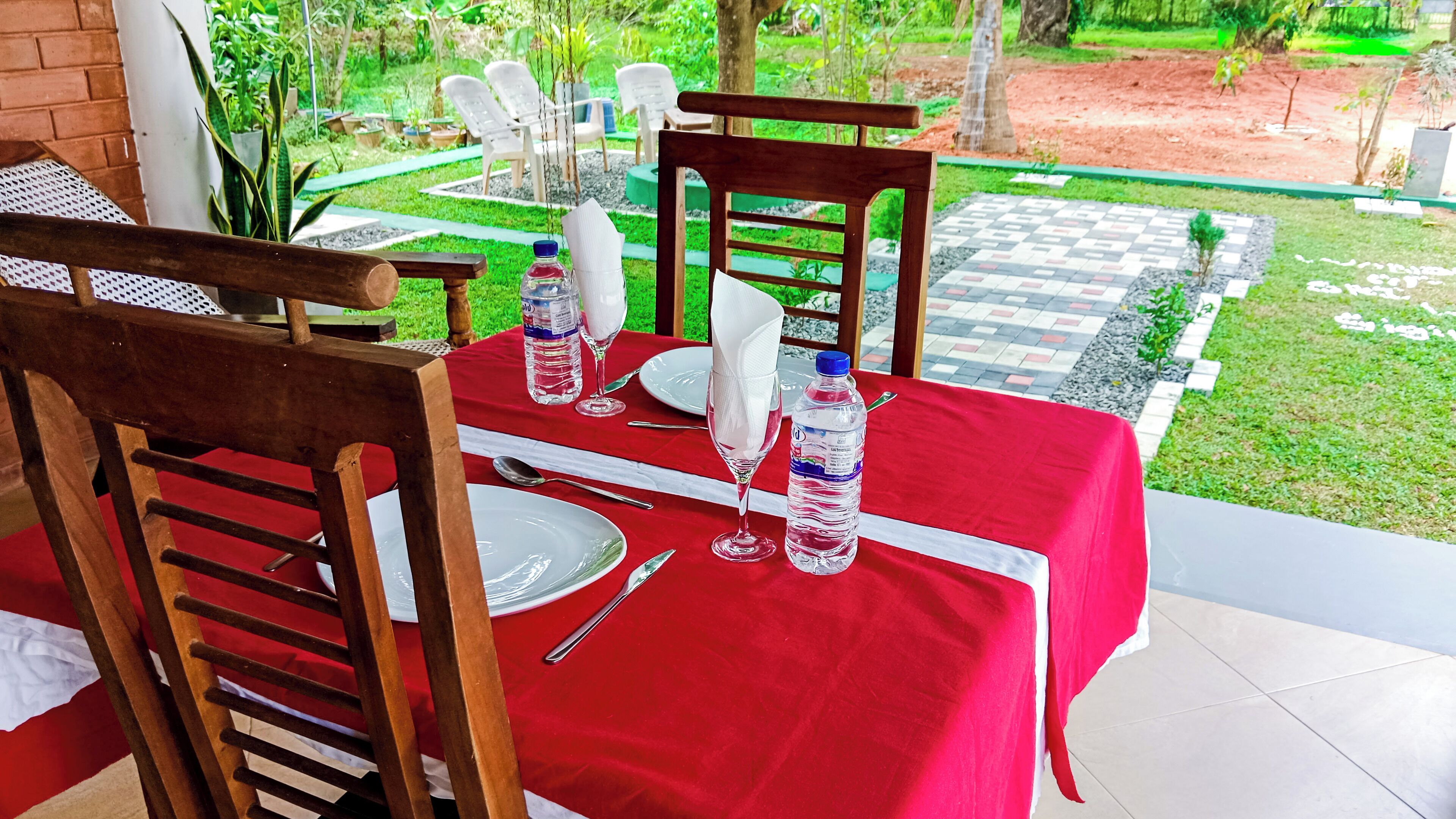 Mesa al aire libre con mantel rojo, sillas de madera y vista a un jardín.