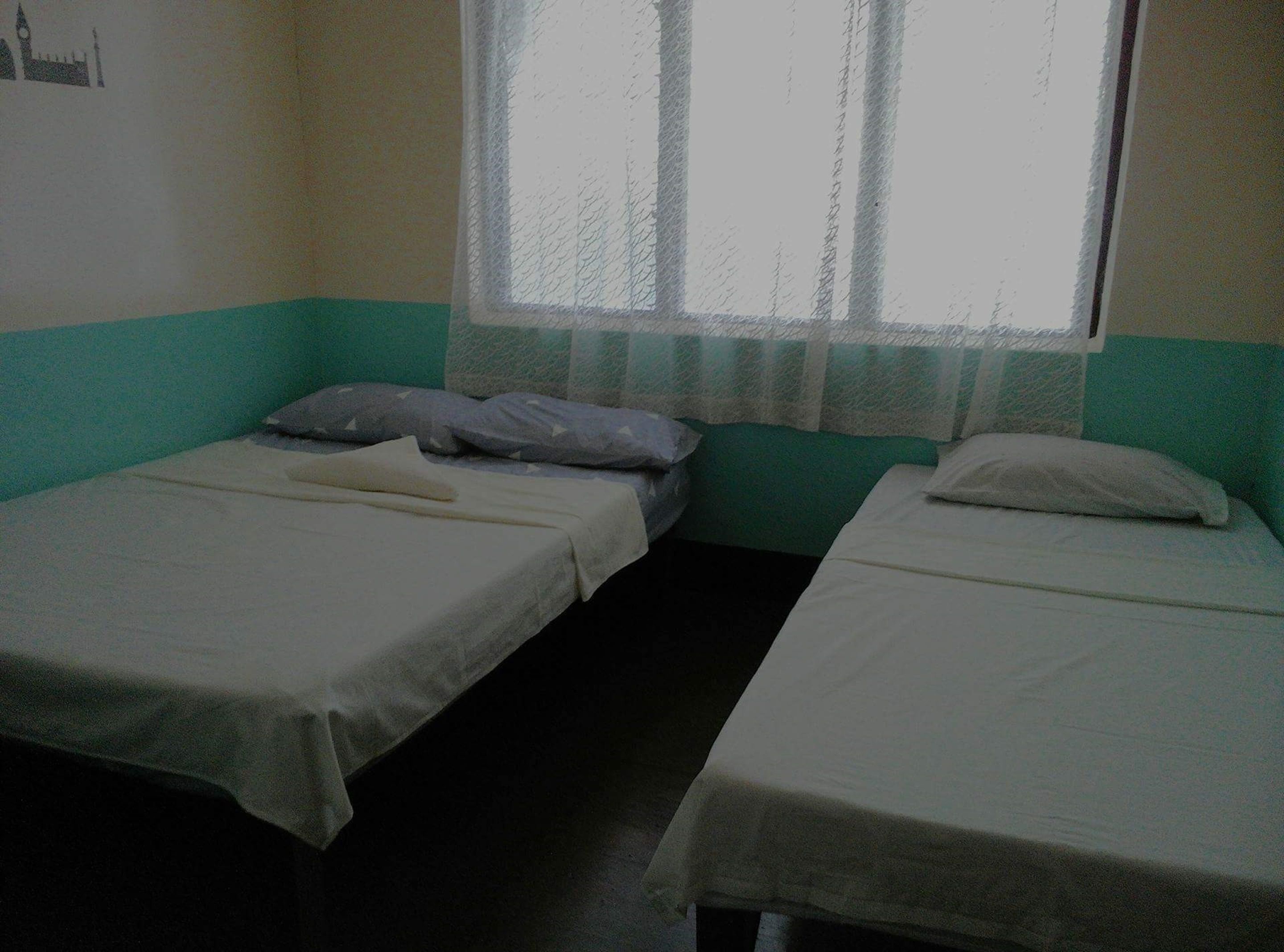 A room with two single beds, a window with a sheer curtain, and a wall with a partial view of a building.