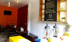 Een restaurant met een schoolbord waarop diverse fruitsappen staan vermeld, een gele tafel en een toonbank met fruit en een blender.